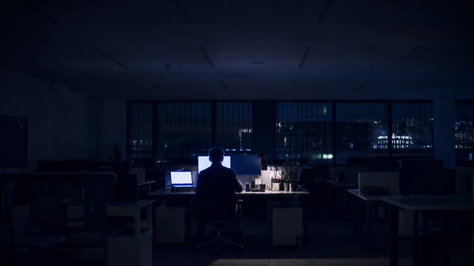 A dark, empty open-plan office late at night with a single workstation lit by monitor glow, one person working alone — the dormant insider who acts selectively and stays below the alarm threshold